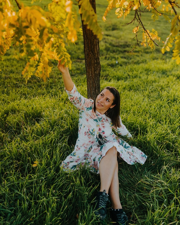 Magnifique femme brune joyeuse assise sous un arbre dans une prairie herbeuse