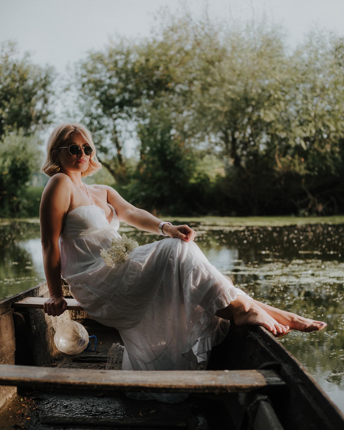 Mireasa într-o rochie de mireasă albă în stil rustic stă într-o barcă pe apă