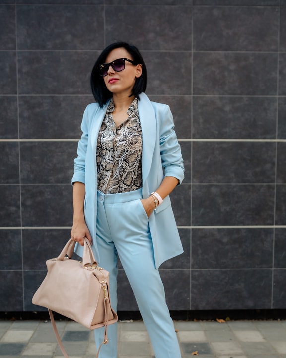 Atemberaubend schöne Geschäftsfrau posiert in blauem Mantel und Hose, während sie eine beige Lederhandtasche in der Hand hält