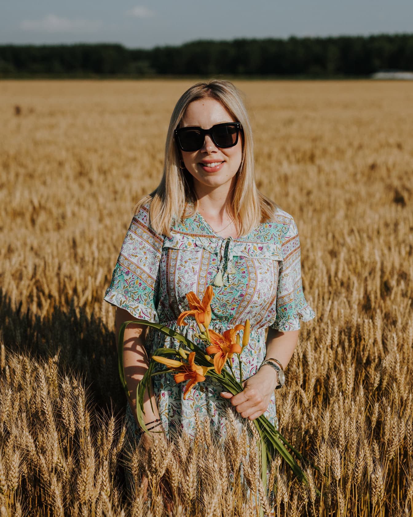 Portret nasmiješene zapanjujuće lijepe žene koja ljeti drži cvijeće ljiljana u polju pšenice