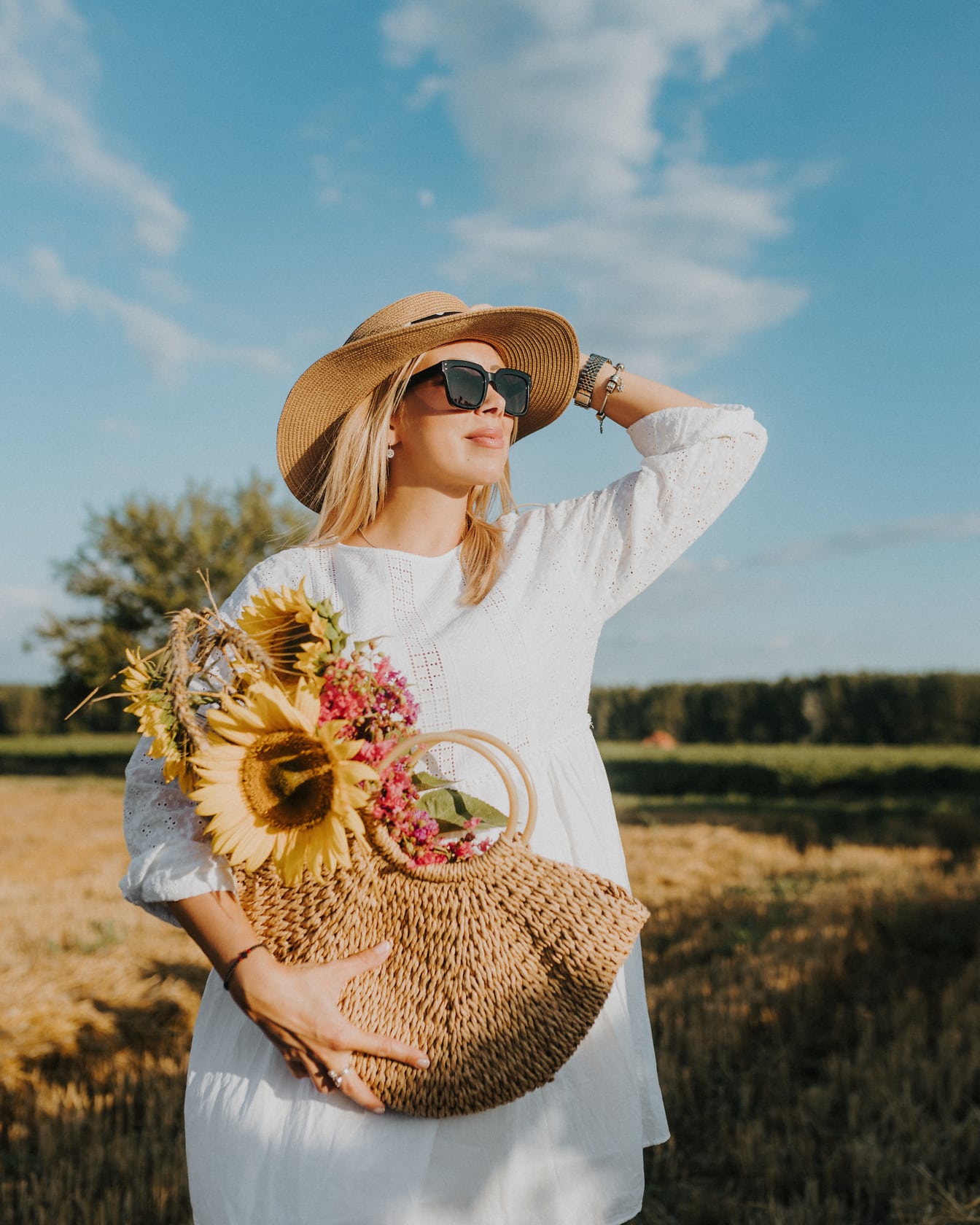 Porträtt av den fantastiskt vackra cowgirl som njuter av att sola på ett fält i en vit klänning och en stråhatt medan du håller en flätad korg med solrosor
