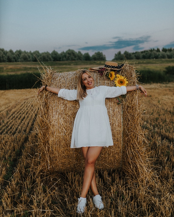 Stunningly beautiful young woman posing in a country style white dress leaning on a haystack in a field of hay