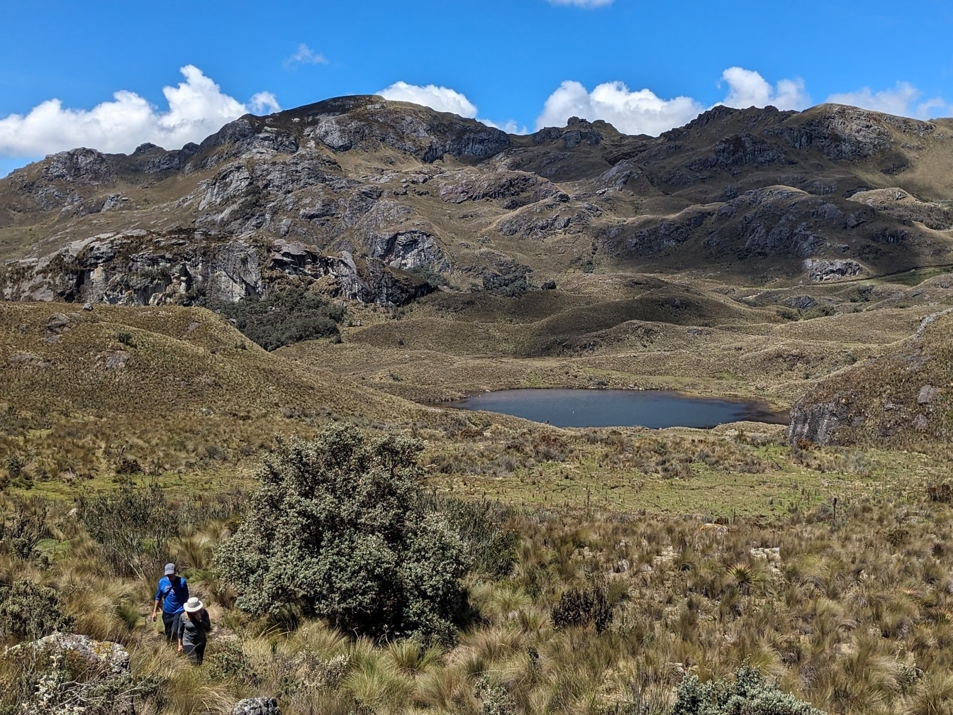 Két túrázó sétál egy füves dombon, háttérben egy tóval az ecuadori Cajes Nemzeti Parkban