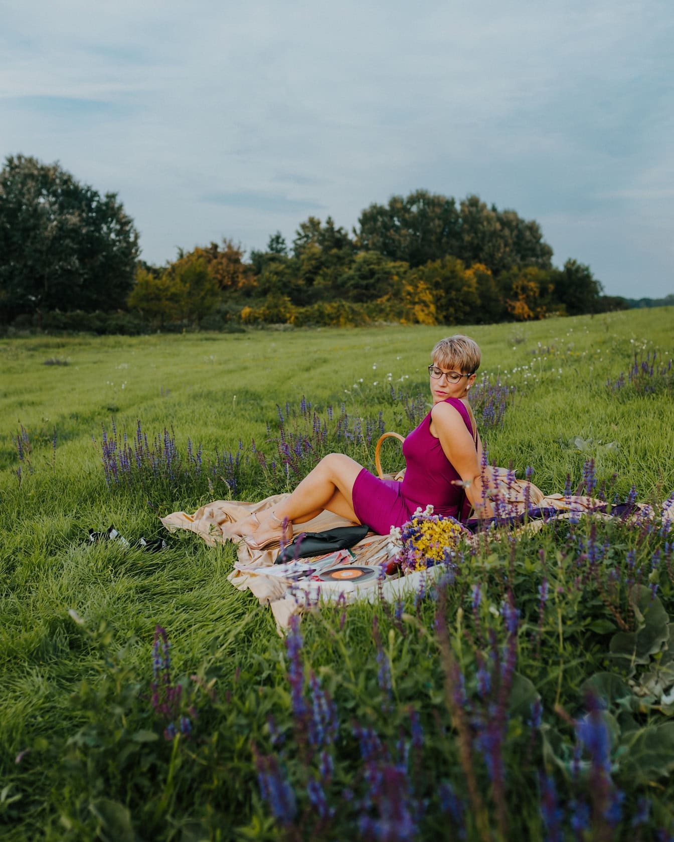 짙은 분홍색 드레스를 입은 여자가 꽃밭에서 피크닉 담요에 앉아 포즈를 취하고 있습니다.