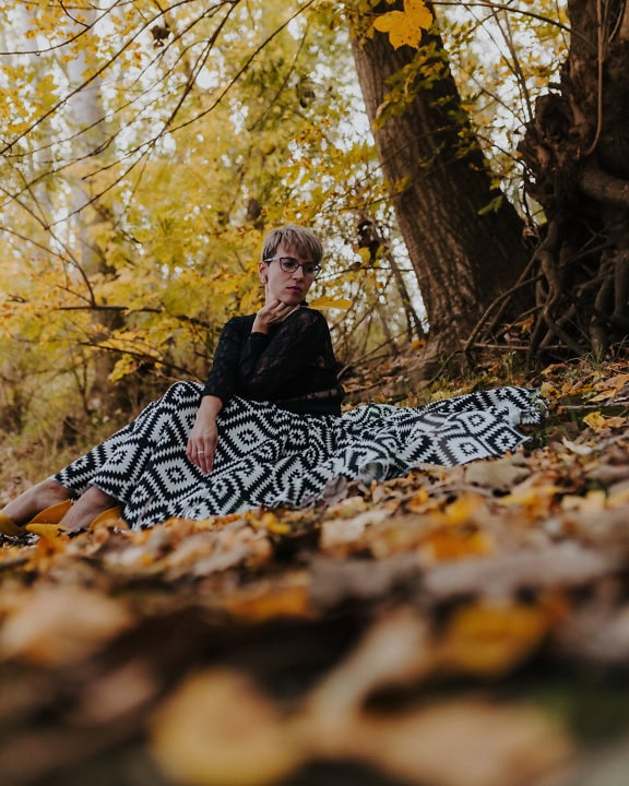 Frau sitzt in einem schwarz-weißen Rock unter einem Baum im Wald im Herbst