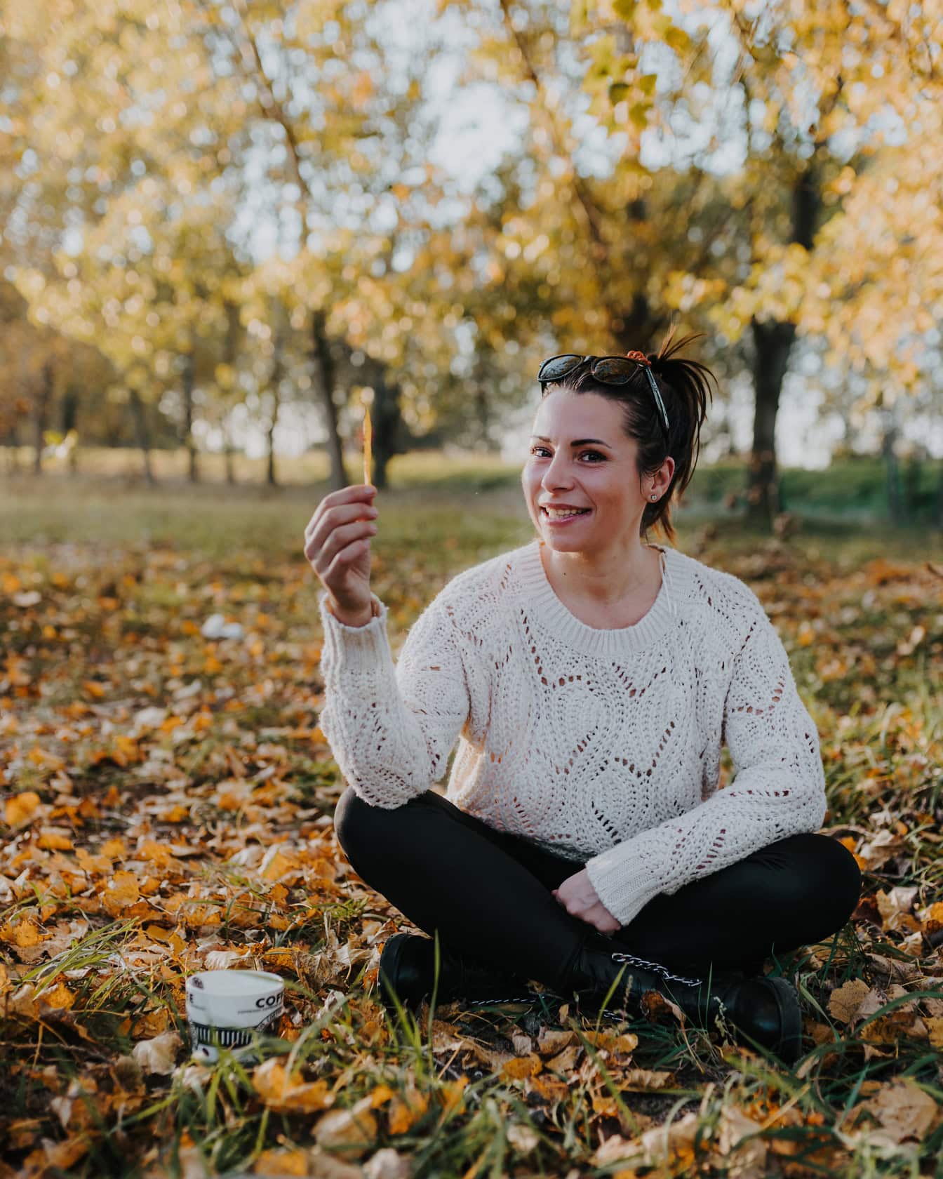 Portré egy mosolygós nő fehér pulóverben, aki a földön ül egy csésze kávéval a parkban ősszel