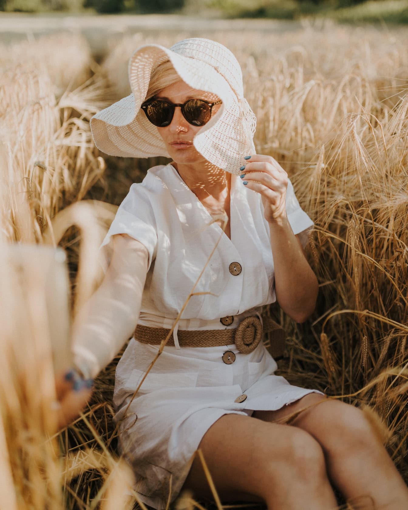 Женщина в белом платье и шляпе сидит на пшеничном поле в солнечный летний день и делает автопортрет на свой мобильный телефон