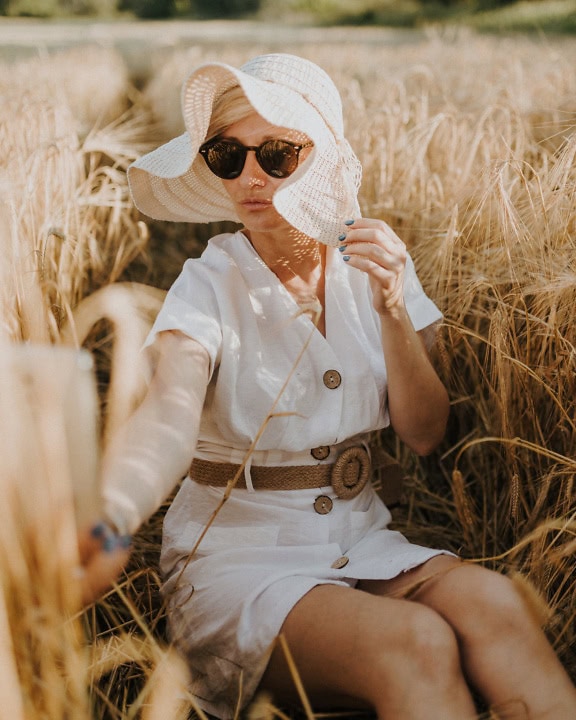 Una donna in un vestito bianco e cappello si siede in un campo di grano in una soleggiata giornata estiva e scatta un autoritratto con il suo cellulare