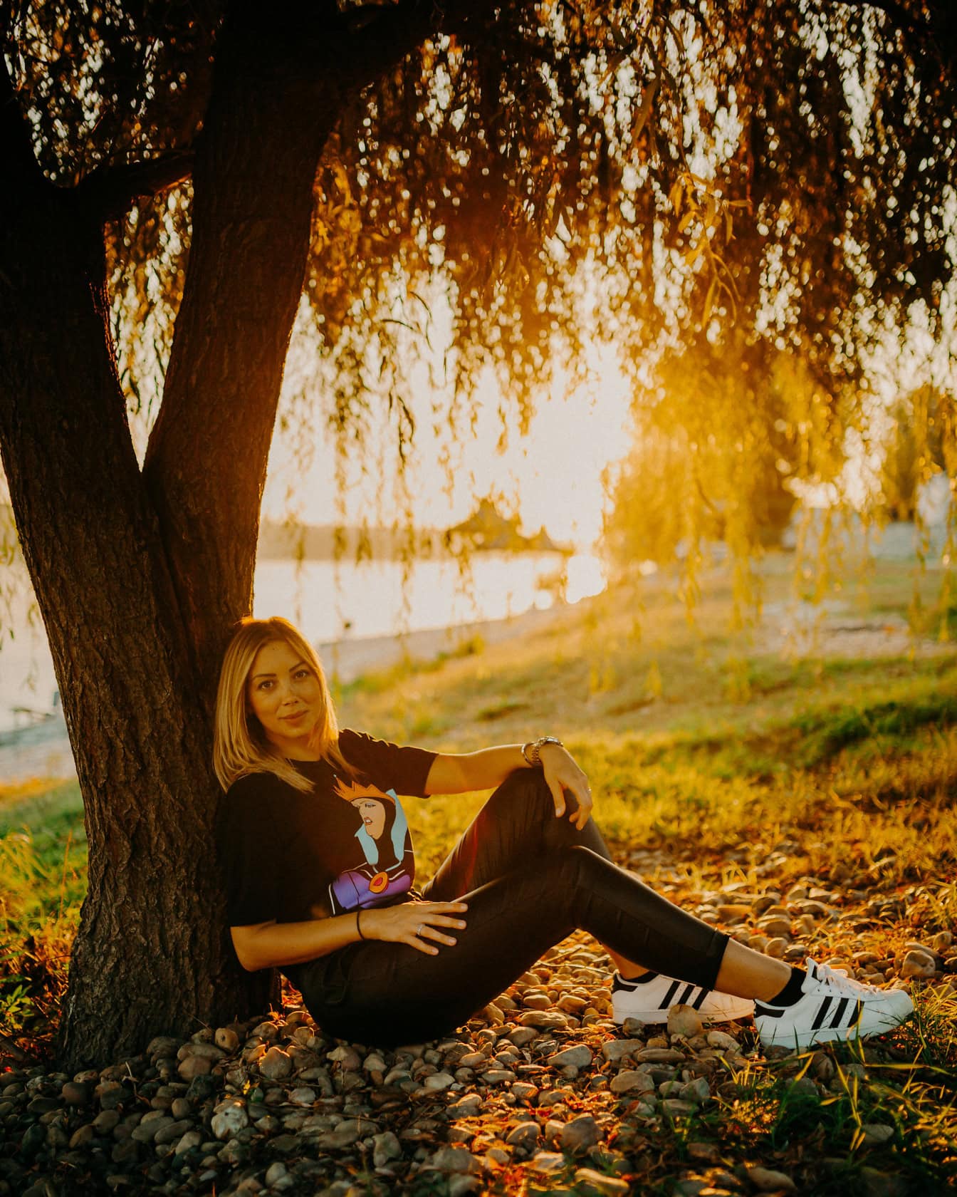 สาวสวยวัยรุ่นนั่งอยู่ใต้ต้นไม้ที่มีพระอาทิตย์ตกที่สวยงามเป็นฉากหลัง