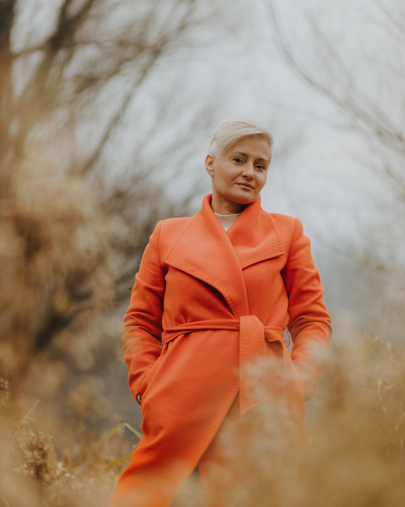 Kvinne med kort blond frisyre som poserer i oransje høstfrakk