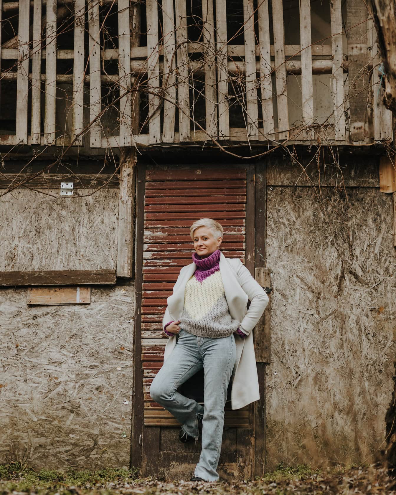 Một người phụ nữ tạo dáng trong quần tây, áo len đan tay và áo khoác trắng dựa vào cửa một nhà kho cũ