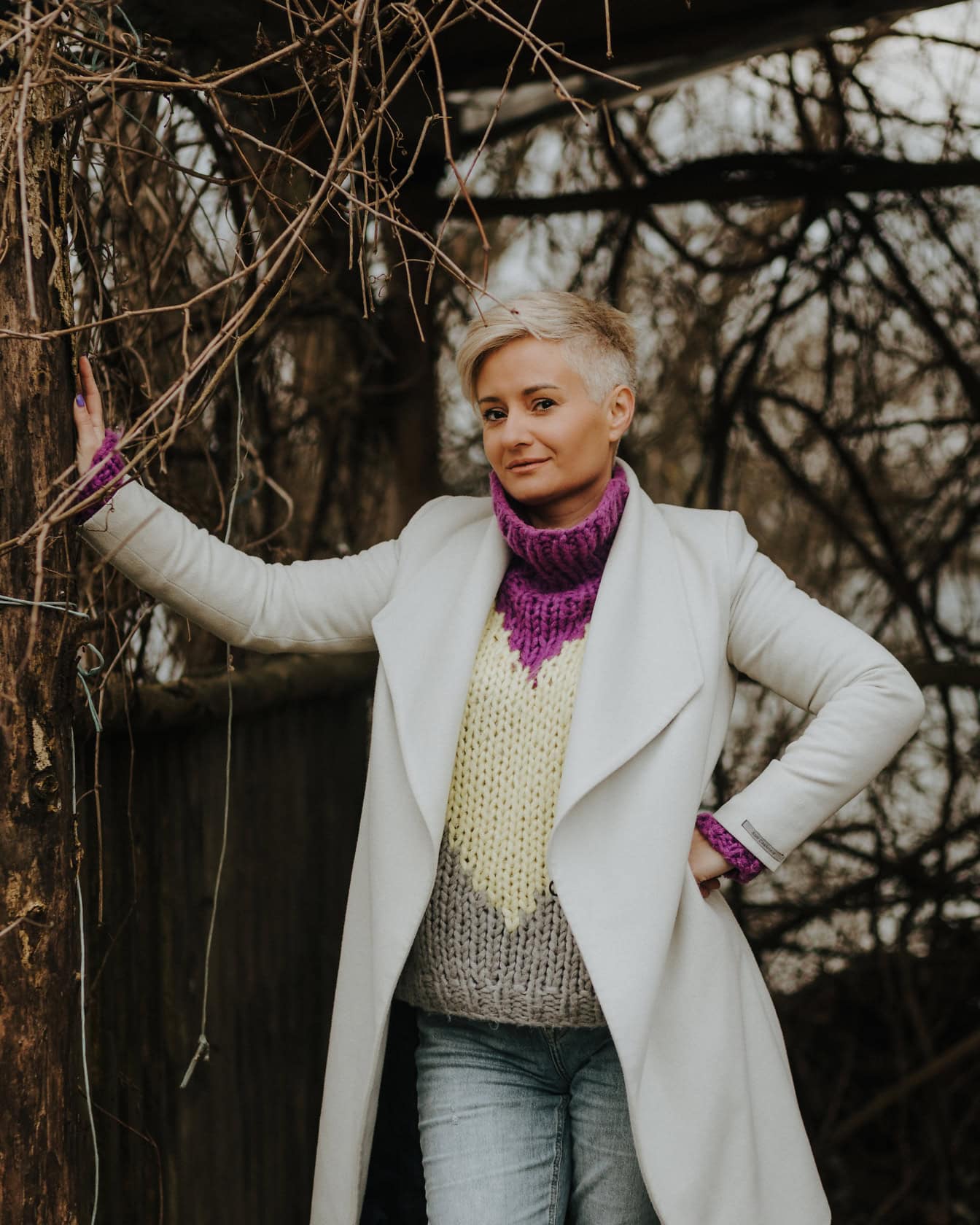 Pretty woman with short hairstyle in a white coat and purple handmade sweater posing leaning on a tree