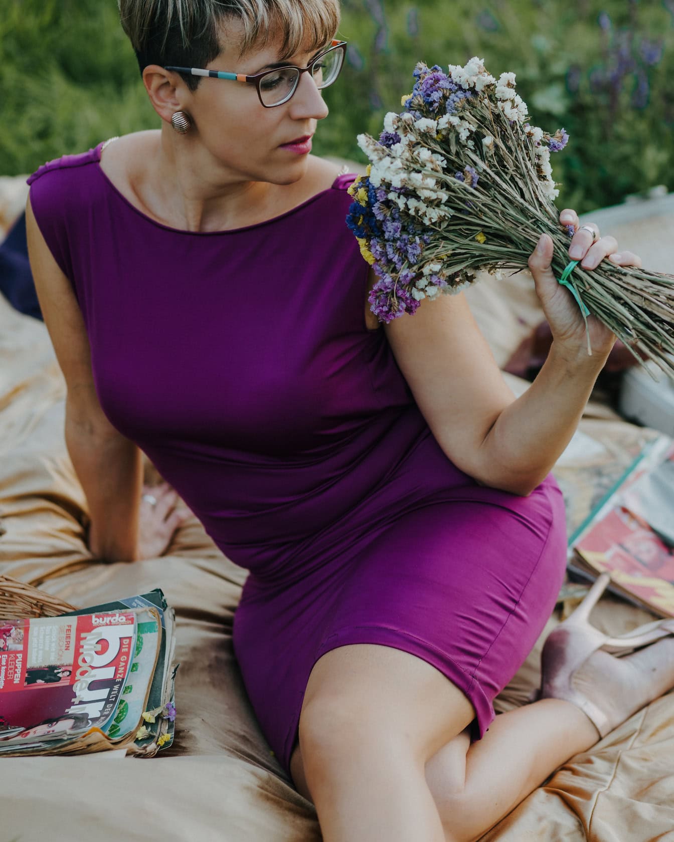 Jóképű nő lila ruhában, kezében csokor mezei virággal, miközben piknik takarón ül
