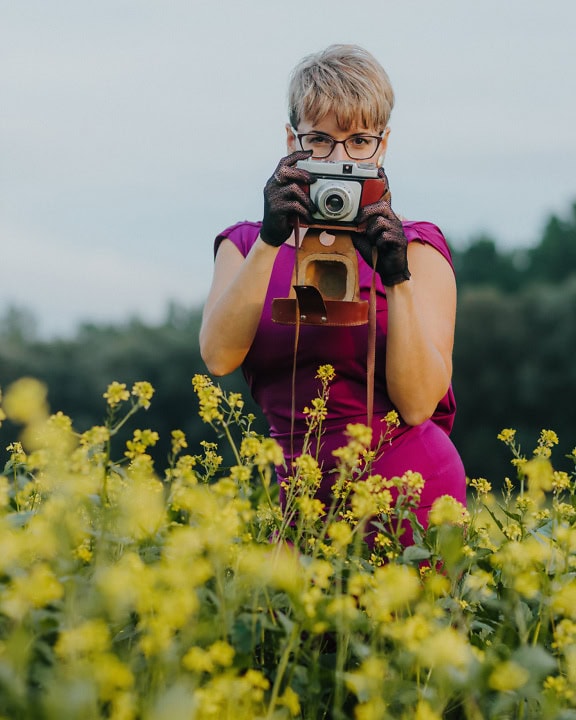 Frau in lila Kleid und Spitzenhandschuhen hält analoge alte Kamera in einem Feld gelber Blumen