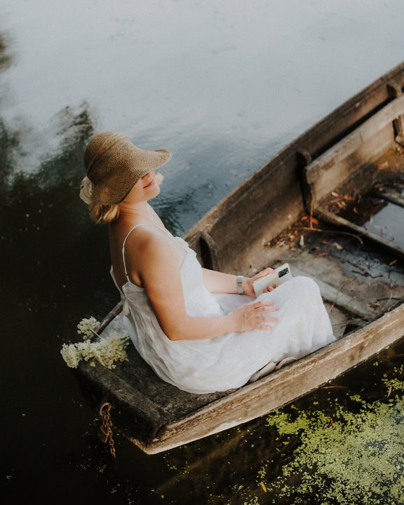 A woman in a white dress and a straw hat sits in an old wooden boat