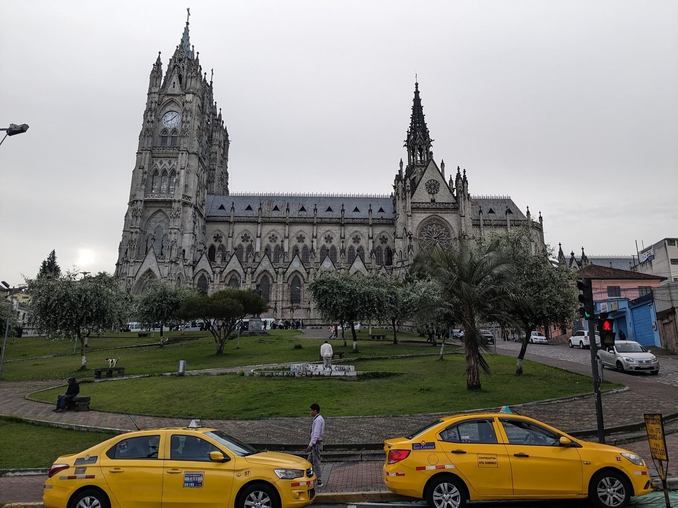 Două taxiuri galbene parcate în fața bazilicii neogotice romano-catolice del Voto Nacional cu un turn cu ceas din Quito, capitala Ecuadorului