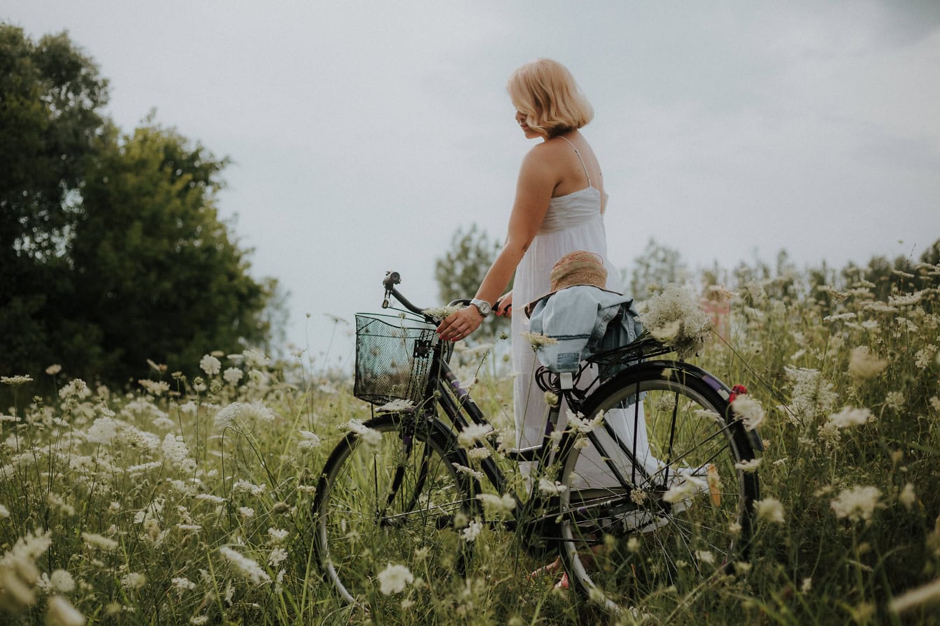 Nainen valkoisessa mekossa polkupyörän kanssa kukkaniityllä kesäpäivänä
