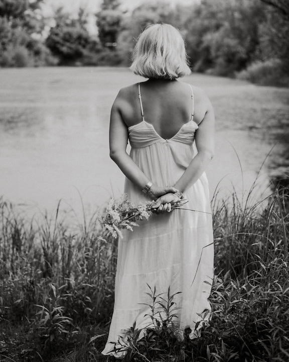 Portrait en noir et blanc de la mariée face à son dos dans une robe blanche dos nu tenant des fleurs