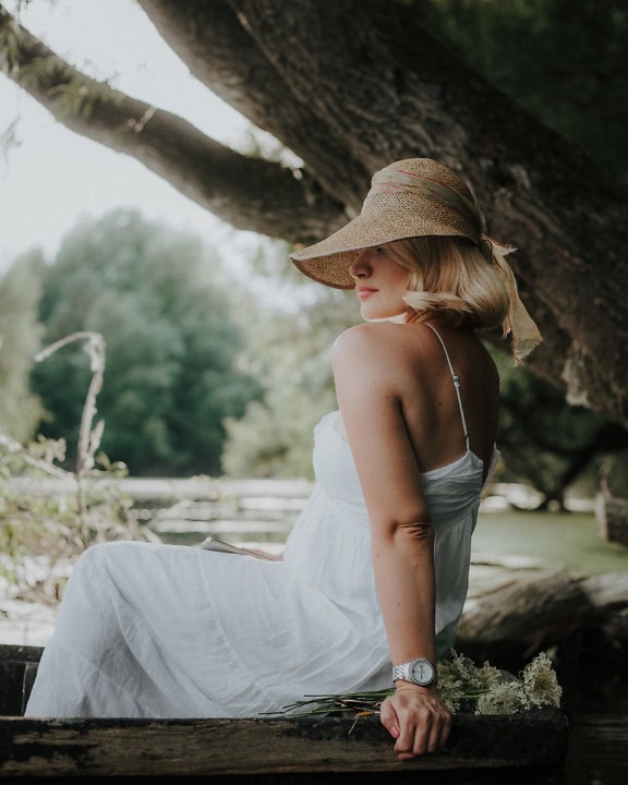 Woman in a elegant backless white dress and hat sitting and posing under a tree