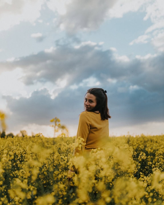 Μια πανέμορφη νεαρή μελαχρινή στέκεται σε ένα ανθισμένο χωράφι με κραμβόσπορο και χαμογελά