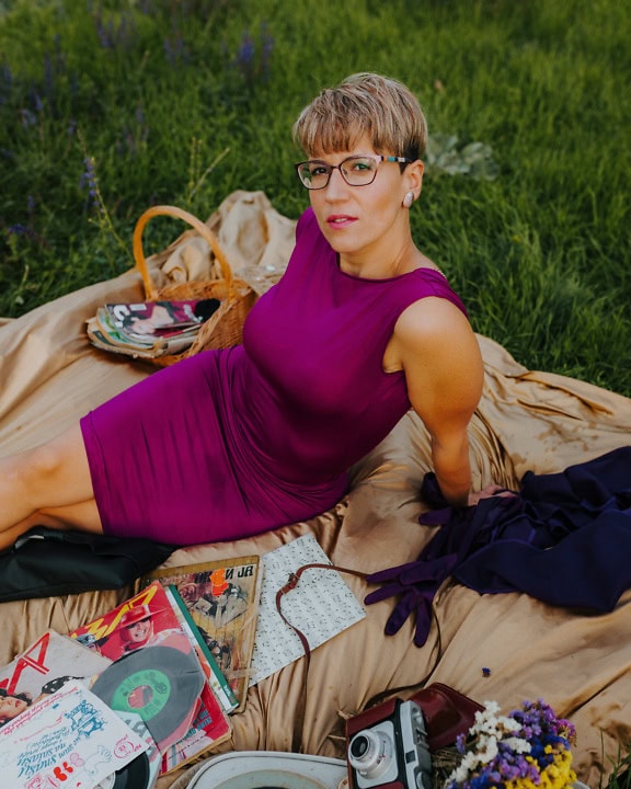 Hübsche blonde Frau in einem lila Seidenkleid, die auf einer Decke sitzt und ein Picknick auf einer Wiese genießt