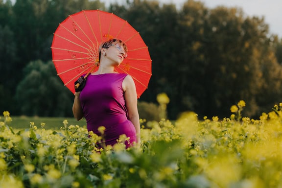 Giovane donna allegra in un vestito viola che tiene un ombrello rosso in un campo di fiori gialli in una soleggiata giornata estiva