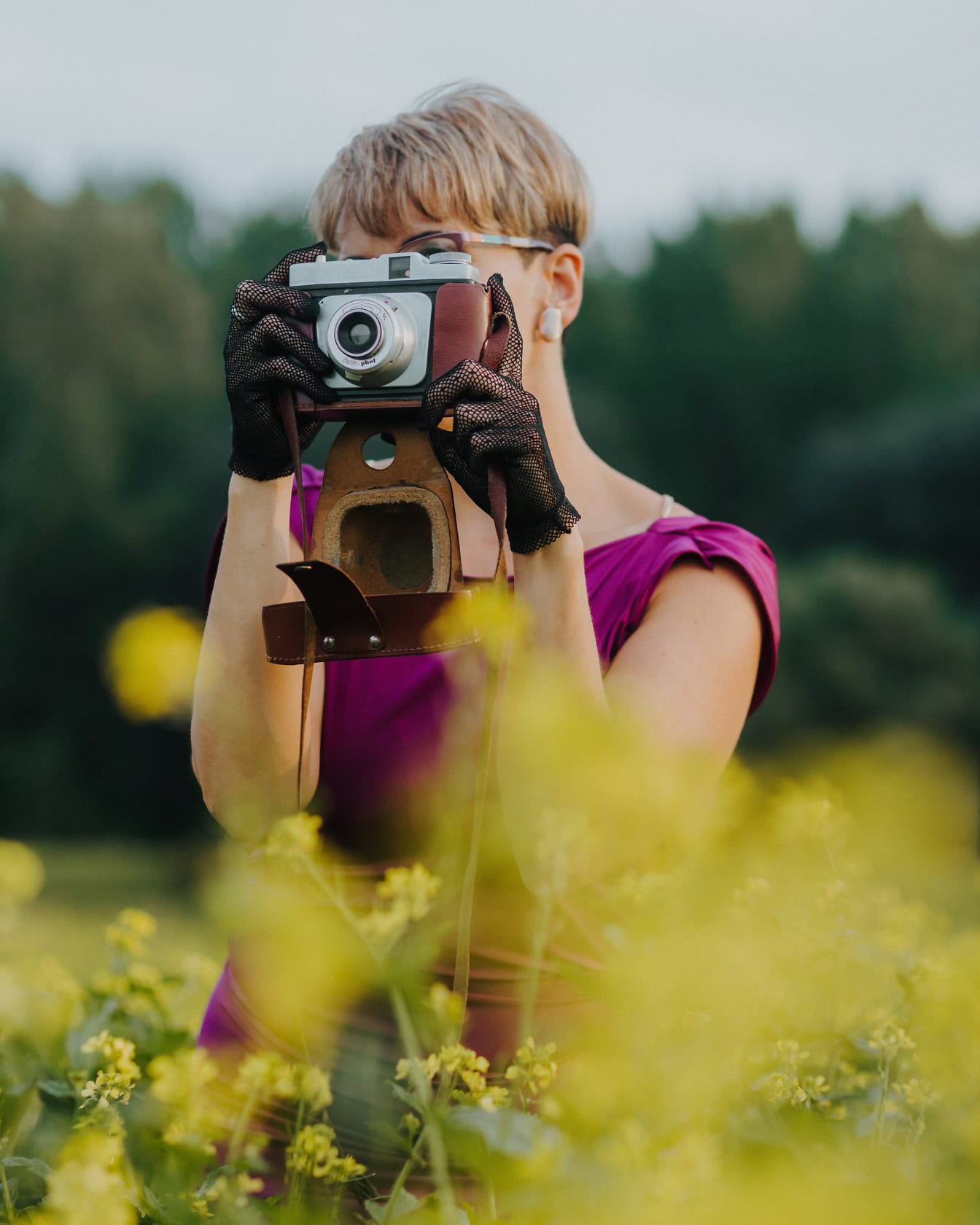 Potret seorang fotografer wanita mengenakan sarung tangan renda dan memegang kamera foto analog tua di depan wajahnya di ladang bunga