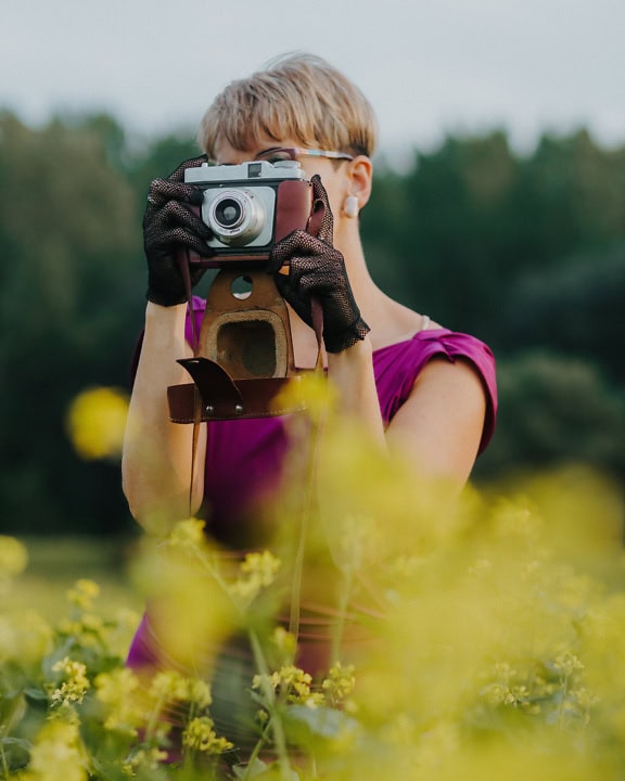 Porträt einer Fotografin, die Spitzenhandschuhe trägt und in einem Blumenfeld eine alte analoge Fotokamera vor das Gesicht hält