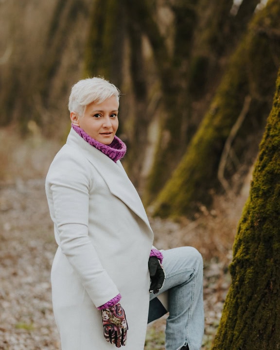 A woman white short blonde hair in a white coat and purple knitted sweater poses for a picture while leaning her foot against a tree