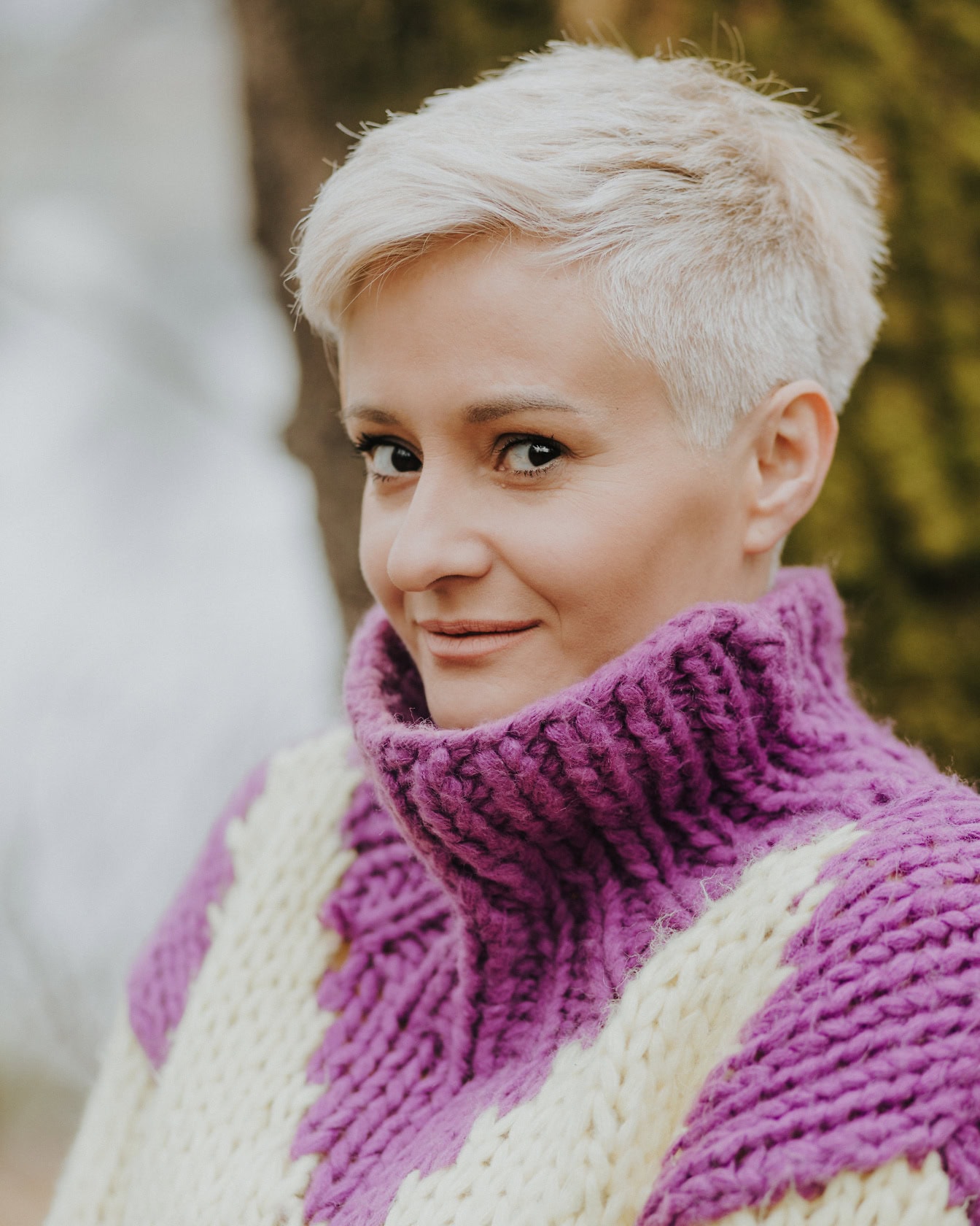 Potret close-up seorang wanita dengan rambut pirang muda pendek mengenakan sweter rajutan buatan tangan ungu-putih