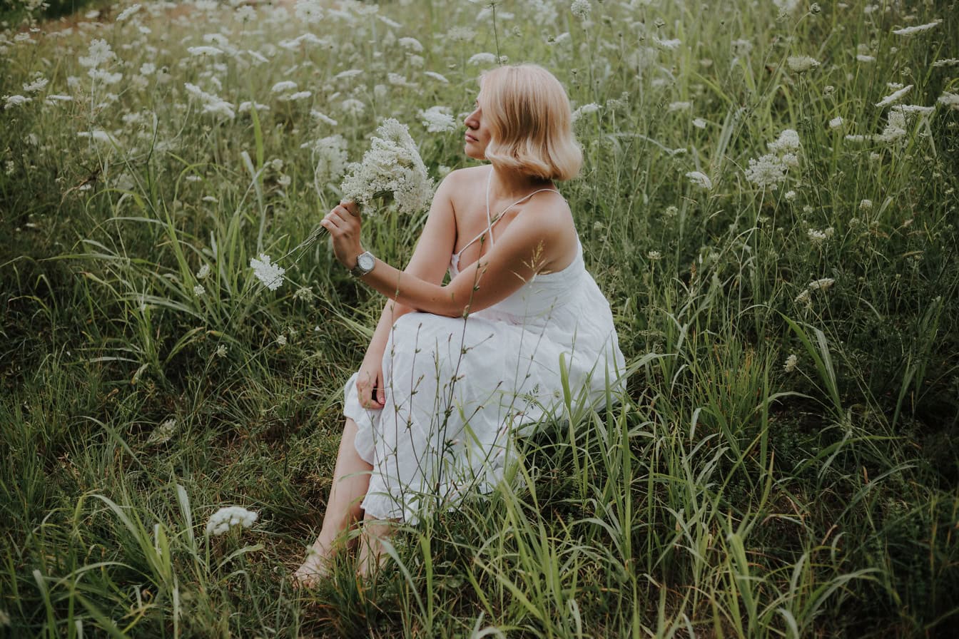 Porträtt av en ung kvinna som sitter i ett fält av blommor och som håller buketten av vita vildblommor i hennes händer