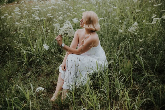 Portræt af en ung kvinde, der sidder på en blomstermark og holder buket hvide vilde blomster i hænderne