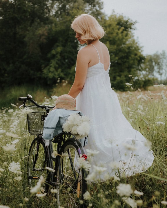 Romantic young woman in a pure white dress with a hat on her bicycle in a field of flowers