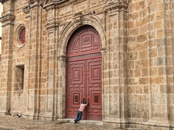 A person praying leaning on the closed front door of the Church of San Pedro Claver in Cartagena, Colombia