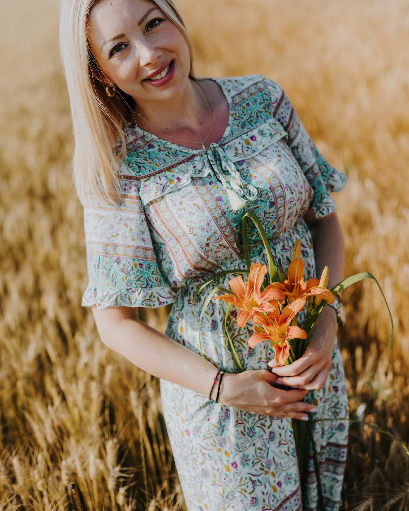 A country young woman in a floral design dress holding a bouquet of orange lily in a wheat field on a warm summer day