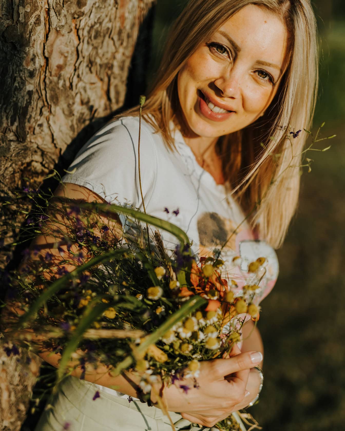 Retrato de una mujer rubia sonriente apoyada en un árbol mientras sostiene un ramo de flores silvestres