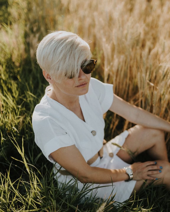 Blonďatá žena s krátkým moderním účesem sedící v bílých šatech v pšeničném poli