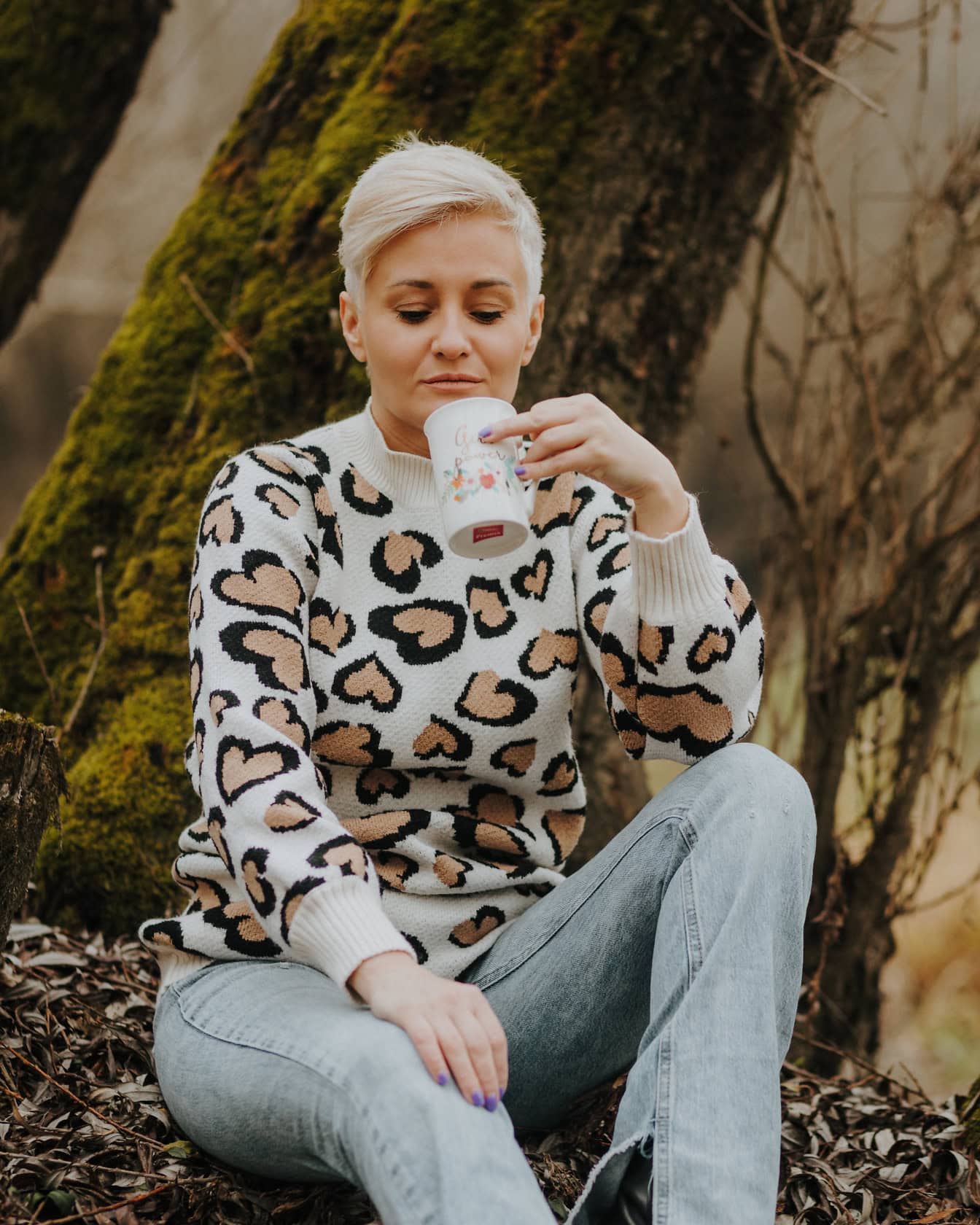 Vrouw met kort blond haar zittend op een boomstronk in sweater en gebleekte broek en en het drinken van een mok