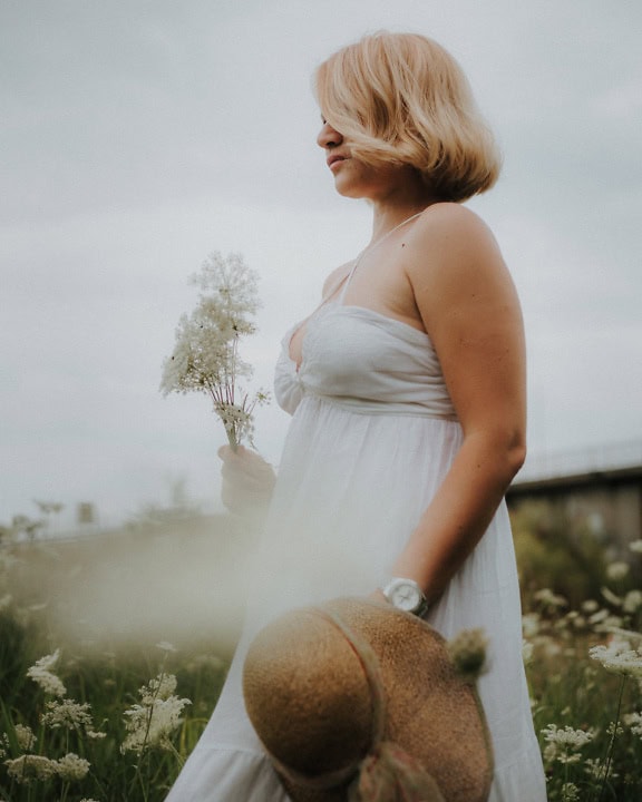 Mujer en un vestido blanco de estilo campestre sosteniendo una flor en un prado