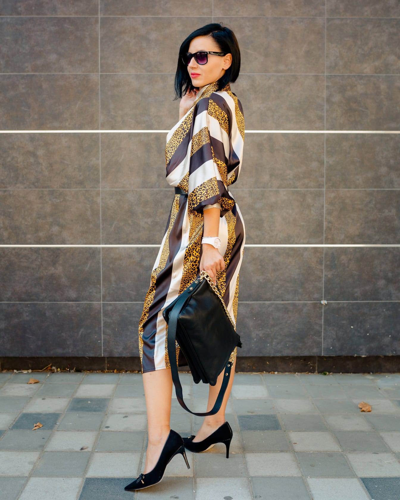 Forretningskvinde poserer i en gylden og brunstribet kjole og med sorte sandaler og læderhåndtaske