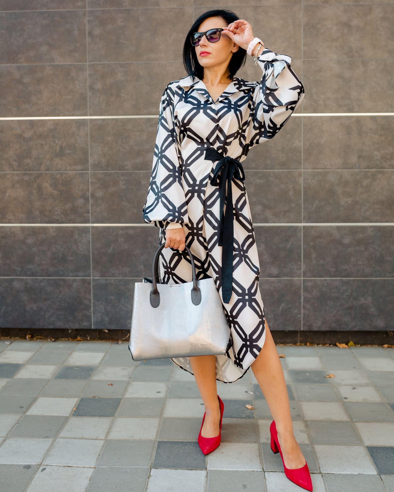 Pretty young woman posing in a black and white dress and red sandals while holding sunglasses in one hand and grayish handbag in another