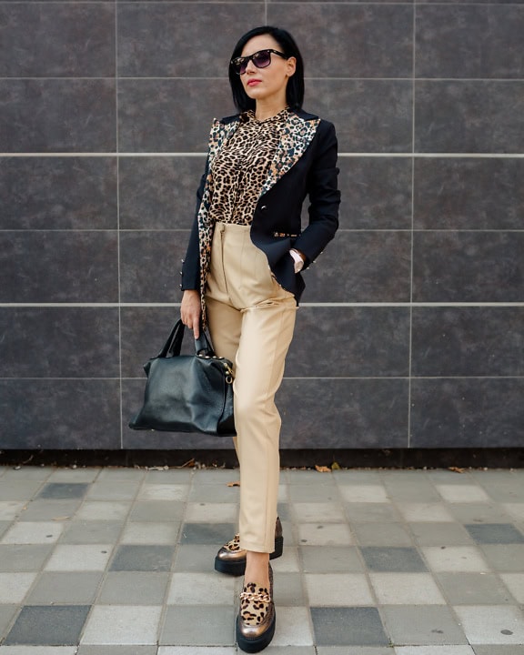 Sebavedomá podnikateľka pózujúca v košeli s leopardou potlačou a kabáte a žltkastých nohaviciach, pričom drží čiernu koženú kabelku