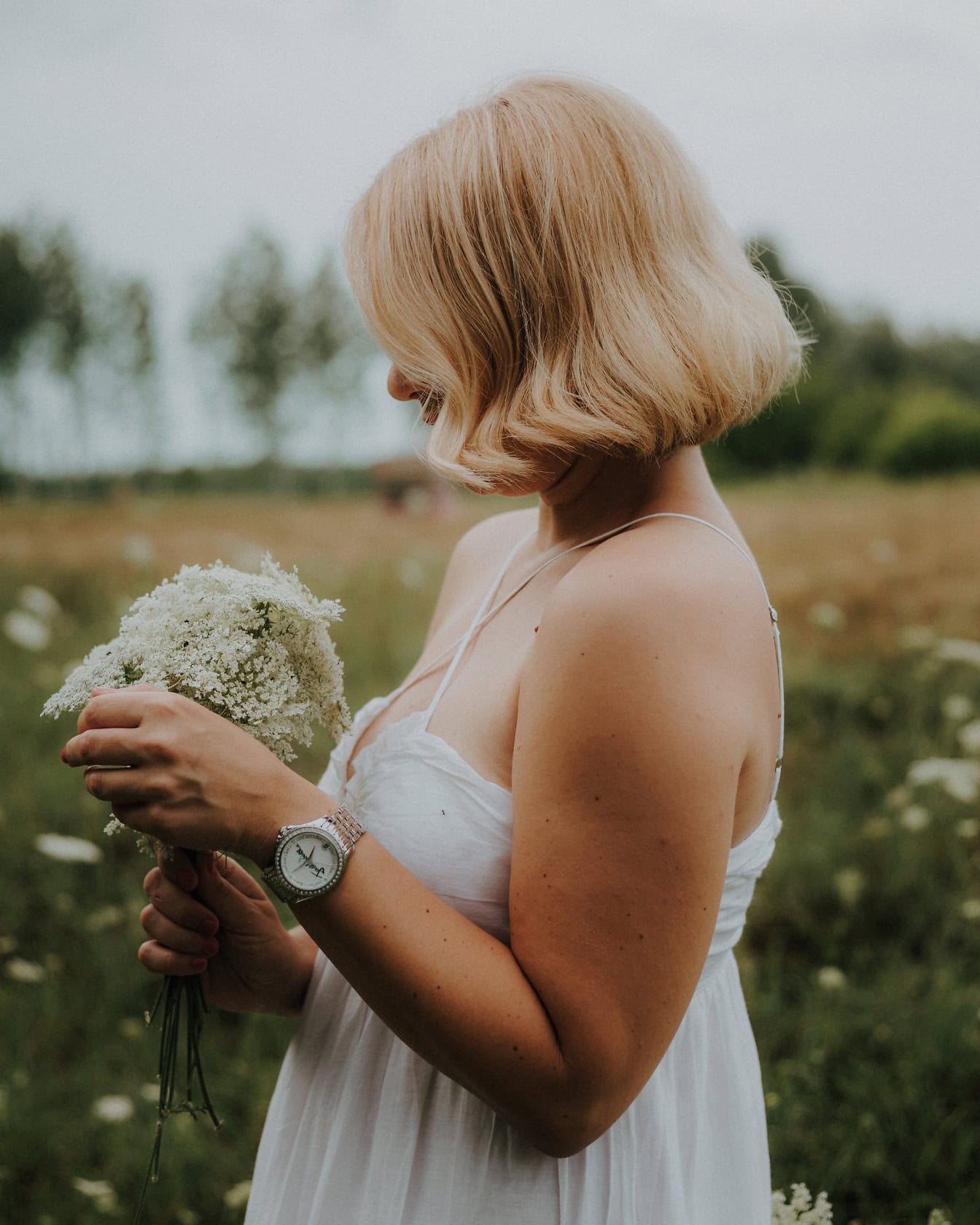 Bočný pohľad na portrét blondínky v bielych šatách vo vidieckom štýle, ktorá drží kyticu bielych poľných kvetov