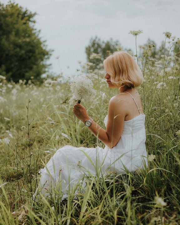 เจ้าสาวผมบลอนด์หน้าตาดีในชุดแต่งงานสไตล์คันทรีนั่งอยู่ในทุ่งหญ้าและเก็บดอกไม้ป่าสีขาว