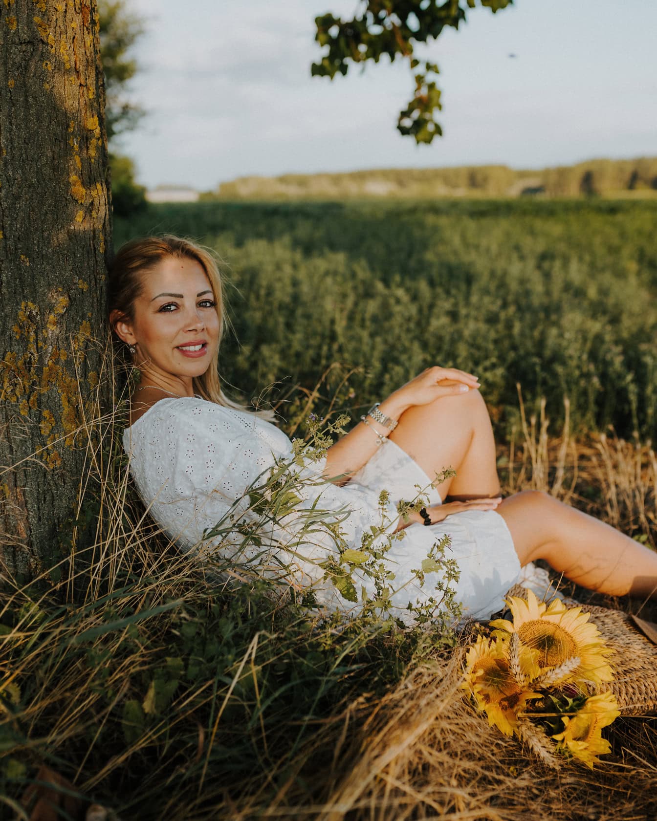 Портрет на зашеметяващо красива блондинка, седнала на тревистата поляна под едно дърво