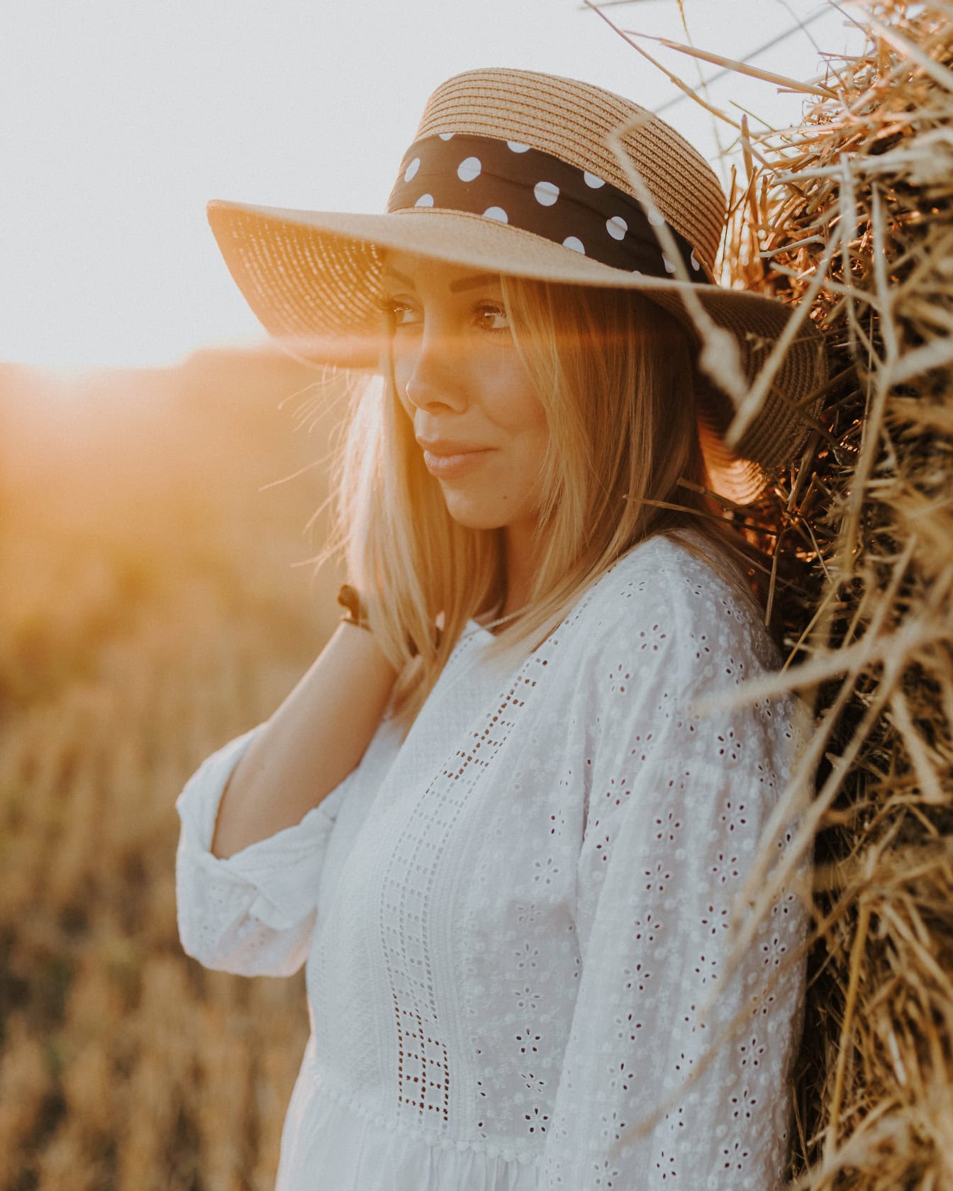 Portrét krásnej blond cowgirl v bielych vidieckych šatách a slamenom klobúku pri opaľovaní na pšeničnom poli opierajúcom sa o kopu sena