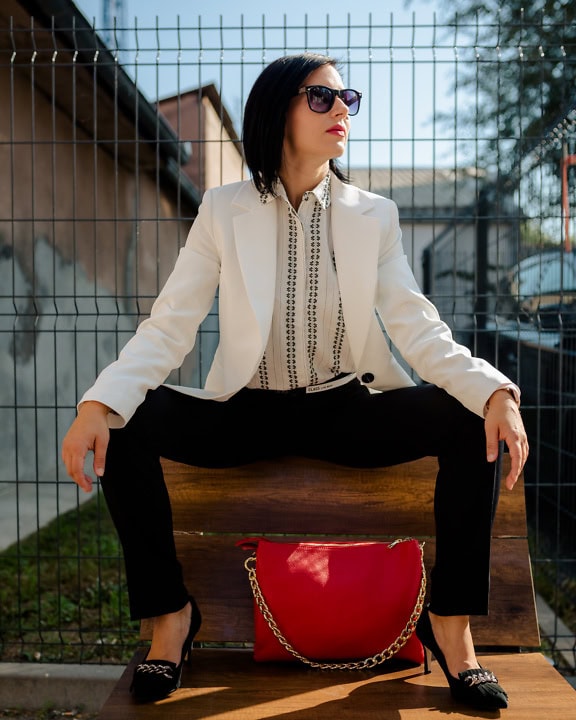 一名年轻女子穿着白色商务外套和黑色裤子，双腿张开坐在长凳上