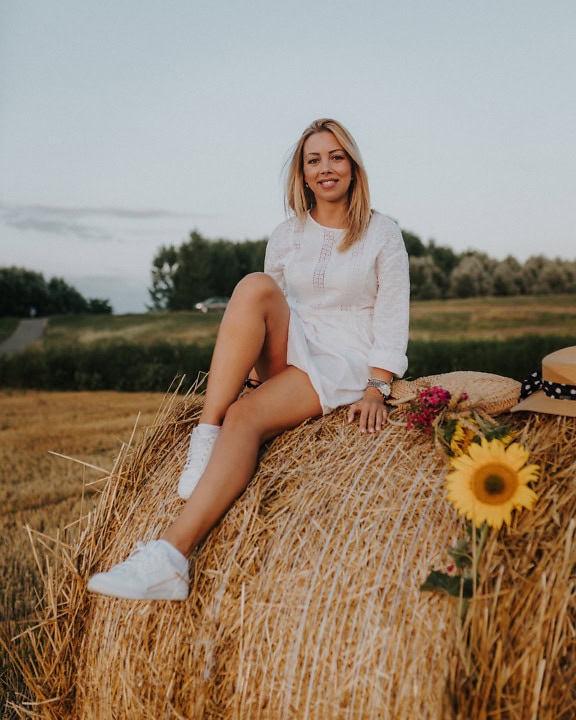 Gadis pirang cantik yang tersenyum duduk di tumpukan jerami dengan gaun pedesaan putih