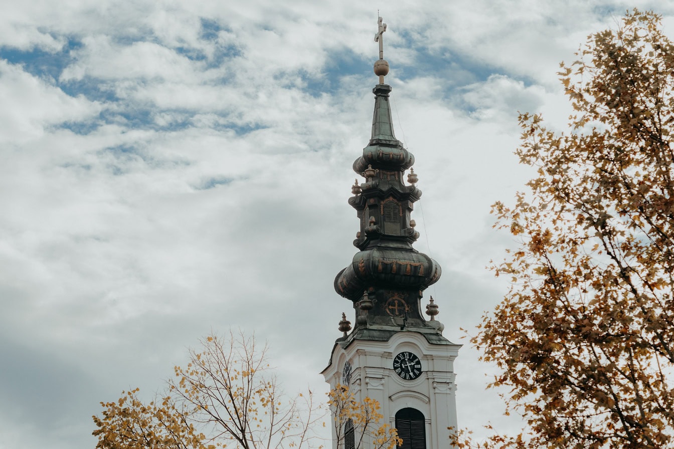 Die serbisch-orthodoxe Kirche des Hl. Johannes des Täufers in der Stadt Bačka Palanka mit einem Uhrenturm und Bäumen und Wolken als Hintergrund