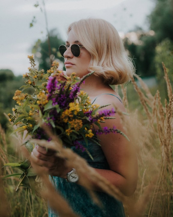 Портрет на жена със слънчеви очила в стил на Джон Ленън, докато държи букет прясно набрани диви цветя в поле